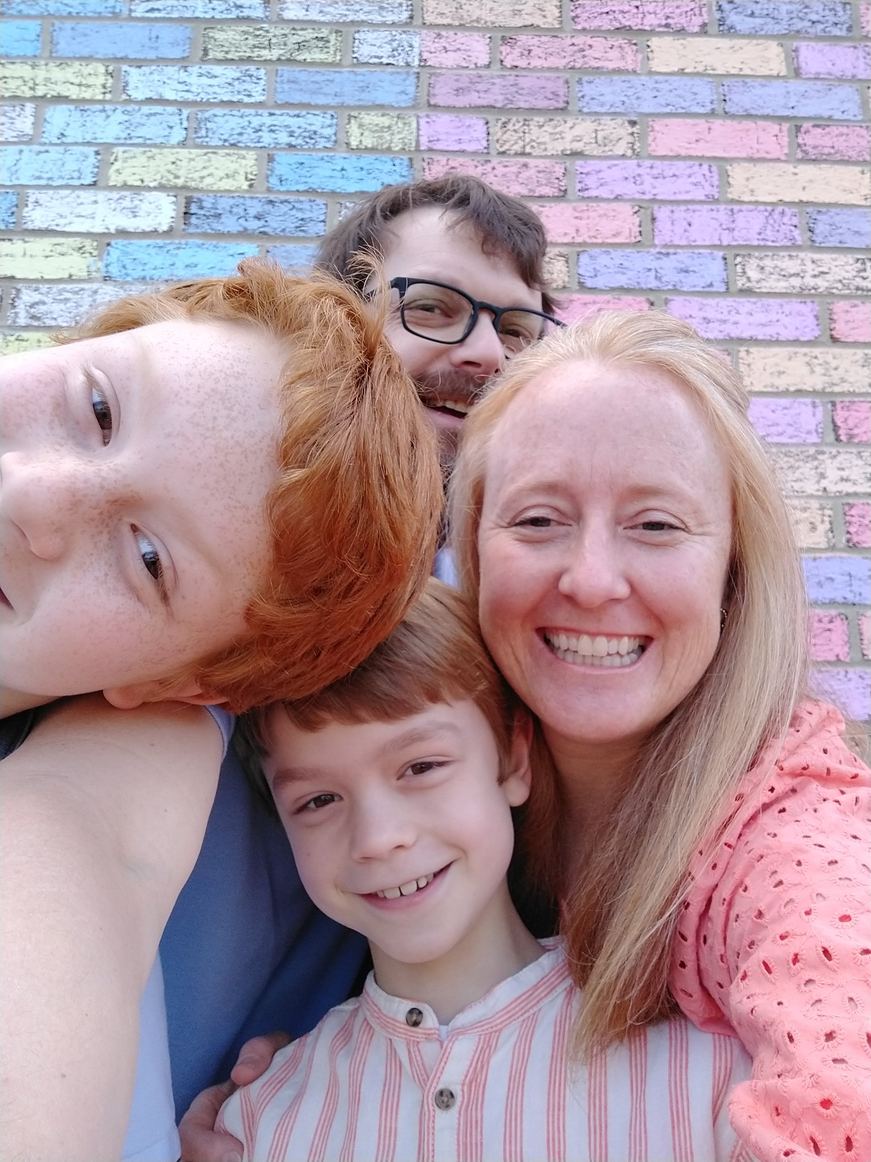 Family of four being silly with pastel brick background and bright clothing. 